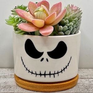 May include: A white ceramic planter with a black Jack Skellington face design, featuring a stitched mouth and eyes. The planter is filled with a variety of succulents, including a pink and yellow succulent in the center.