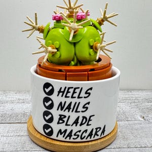 May include: A whimsical cactus sculpture made of green and tan blocks, topped with pink accents, sits in a white ceramic pot. The pot features a checklist with the words: Heels, Nails, Blade, and Mascara.