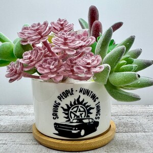 May include: A white ceramic planter with the text "Saving People, Hunting Things" and a black graphic of a car and a symbol. The planter holds faux succulents in shades of green, pink, and burgundy. It sits on a small, round wooden coaster.
