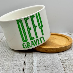 May include: White ceramic planter with a green "DEFY GRAVITY" graphic. The planter has a matching wooden base.