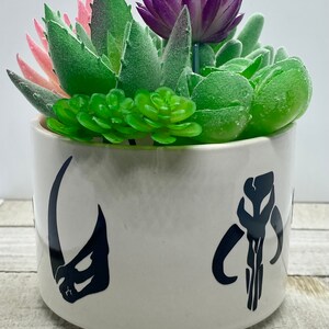 May include: White ceramic planter with a black Mandalorian symbol and a black symbol of the character Grogu from the Star Wars series. The planter is filled with green and pink artificial succulents.