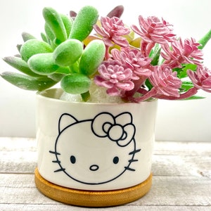 May include: A white ceramic planter with a black Hello Kitty design, featuring a pink bow and whiskers. The planter is filled with green and pink artificial succulents.