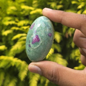 May include: An oval-shaped gemstone with a green base color and vibrant pinkish-red inclusions. The polished stone is held between fingers against a blurred background of green foliage. The stone's surface has a smooth, glossy appearance.