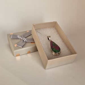 May include: A silver pendant necklace with a teardrop-shaped gemstone, displaying red and green colours, is presented in an open gift box. The box is beige with a silver bow and polka dots. The necklace rests on white foam.