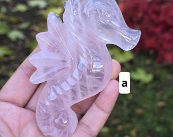 May include: A carved rose quartz seahorse figurine. The sculpture is a pale pink color and features detailed wings, a curved body, and a detailed face. The seahorse is held in a hand against a blurred background of green and red.