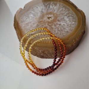 May include: Three strands of amber beads in graduated shades of yellow and brown. The beads are round and smooth, and they are arranged in a circle on a light brown agate slice.