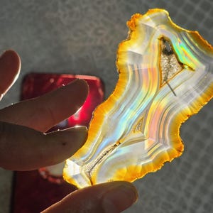 May include: A translucent agate slice, displaying a mix of colours including white, yellow, orange, and iridescent hues. The stone's irregular shape and banded patterns are visible. The agate is held up to the light, showcasing its natural beauty.