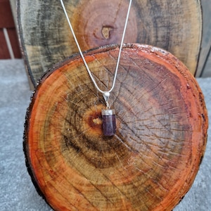 May include: A silver chain necklace with a purple crystal pendant. The pendant is a rectangular prism shape and is hanging on a wooden surface.