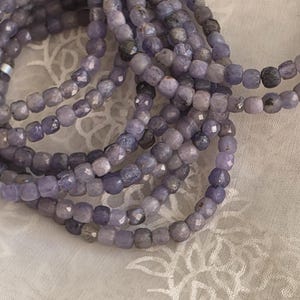 May include: Close-up of multiple strands of lavender-coloured, faceted beads. The beads are small, square-shaped, and strung together, creating a layered effect. The colour varies from light to dark purple, with some grey tones.
