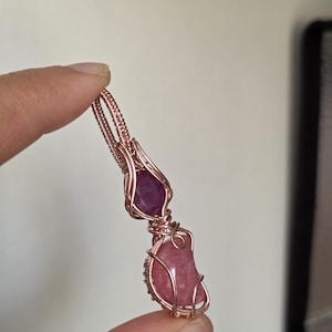 Puede incluir: Un colgante artesanal con dos piedras preciosas naturales envueltas en alambre de cobre. La piedra superior es de color magenta intenso, mientras que la inferior es de un rosa suave. El trabajo de alambre crea un diseño intrincado y abierto, mostrando las formas naturales de las piedras.