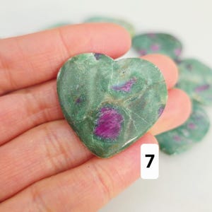 May include: A heart-shaped stone with a green base colour and vibrant magenta inclusions. The stone is held in a hand, with other similar stones visible in the background. The number "7" is visible on a white background.