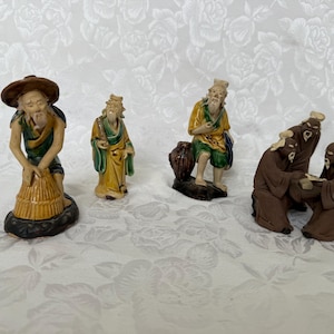 Op de afbeelding: Een verzameling kleine, gedetailleerde beeldjes die oudere figuren in verschillende poses uitbeelden. De beeldjes hebben een reeks kleuren, waaronder bruin, geel, groen en beige. Eén figuur draagt een hoed en werkt met een mand.