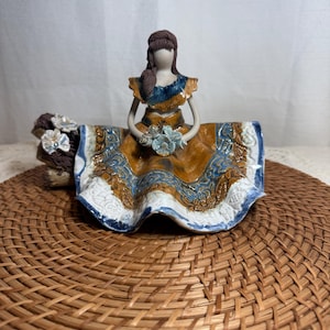 Può includere: Statuina in ceramica di una donna in abito tradizionale, con un bouquet di fiori blu. L'abito presenta motivi marroni, blu e bianchi. La statuina è su un tappetino intrecciato.