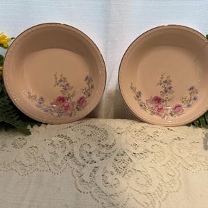 Puede incluir: Dos cuencos de cerámica rosa a juego con diseños florales. Cada cuenco presenta un delicado patrón de flores rosas, moradas y azules. Los bordes tienen un ribete dorado y un borde texturizado. Los cuencos están sobre un mantel de encaje blanco.