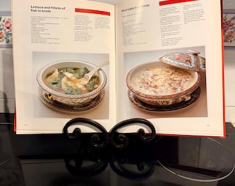 Könnte beinhalten: Ein aufgeschlagenes Kochbuch auf einem schwarzen Metallständer zeigt Rezepte für Salat mit Fischfilets in Brühe und West Lake Rindfleischsuppe. Jede Seite enthält detaillierte Anweisungen und ein Foto des zubereiteten Gerichts.
