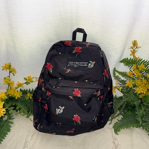 May include: Black Jansport backpack with a floral pattern of red roses and small hummingbirds. The backpack has a top handle and a front pocket. The Jansport logo is visible on the front.