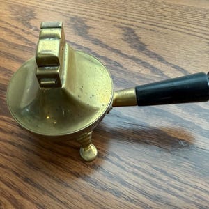 Puede incluir: Una tetera de latón vintage con un mango de madera negro. La tetera tiene un cuerpo redondo, tres patas pequeñas y una tapa con bisagras. El mango está unido al lateral de la tetera y es de madera negra.