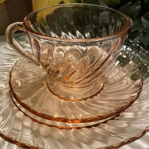 May include: A vintage pink glass teacup and saucer set. The cup and saucer have a swirled design. The set is made of transparent pink glass. The cup sits on a matching saucer, with a second saucer visible below.