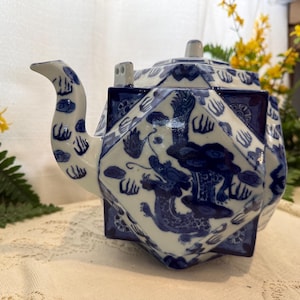 Pode incluir: Uma chaleira de cerâmica branca e azul com um desenho de dragão. A chaleira tem uma forma geométrica com um bico e uma alça. A chaleira é decorada com motivos florais e de dragões azuis. É um artigo decorativo.