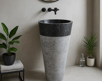 May include: A modern pedestal sink with a black marble top and a gray, textured base. The sink is in a bathroom setting with a black faucet, a round mirror, and potted plants. The overall design is minimalist and contemporary.
