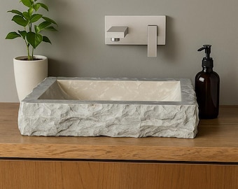 May include: A rectangular stone sink with a textured exterior and a smooth interior. A modern faucet is mounted on the wall above the sink. A brown glass soap dispenser and a potted plant are on the wooden surface.