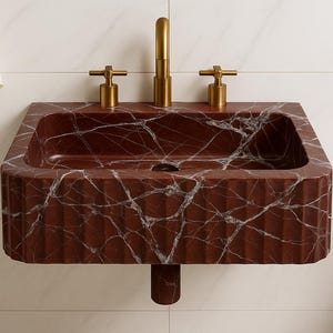May include: A rectangular, wall-mounted sink made of red marble with white veining. The sink has a fluted front and is paired with gold-colored faucets. A white towel hangs on the left.
