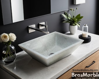 May include: A white marble vessel sink with a rectangular shape and a beveled edge. The sink is installed on a white countertop with a wooden cabinet underneath. A chrome faucet is mounted on the wall behind the sink.
