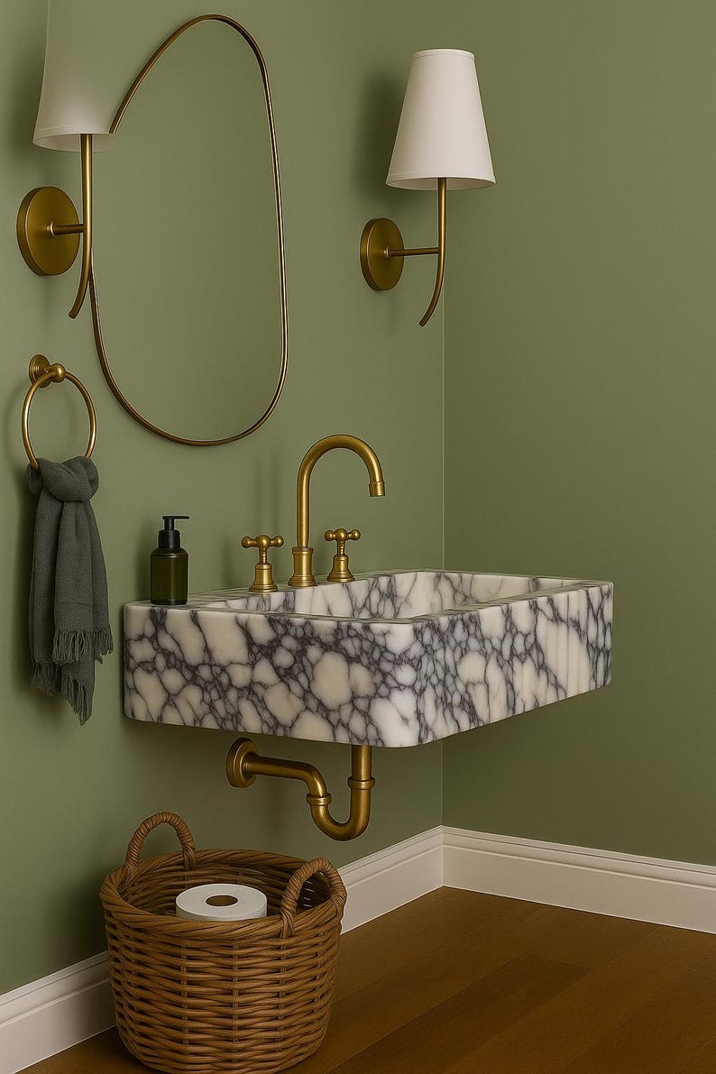Pu&ograve; includere: Un lavandino in marmo con rubinetti dorati &egrave; montato su una parete verde. Uno specchio con cornice dorata e applique con paralumi bianchi sono sulla parete. Sono presenti anche un cestino di vimini e un anello portasciugamani.