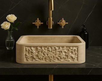 May include: A beige stone rectangular sink with a carved floral pattern. Gold-coloured taps and a faucet are mounted on the wall above. A clear vase with white roses and a black soap dispenser sit on the dark countertop.