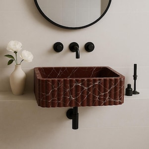 Può includere: Un lavabo rettangolare in marmo rosso con un design scanalato, montato su una parete bianca. Il lavabo ha un rubinetto e accessori neri. Un vaso con fiori bianchi e un candeliere nero sono a sinistra e a destra. Uno specchio nero e rotondo è appeso sopra.