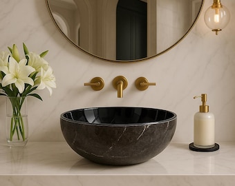 May include: A modern bathroom sink with a dark, marbled finish. The round vessel sink sits on a white countertop, complemented by gold fixtures and a matching soap dispenser. A round mirror and a vase of white lilies complete the elegant design.