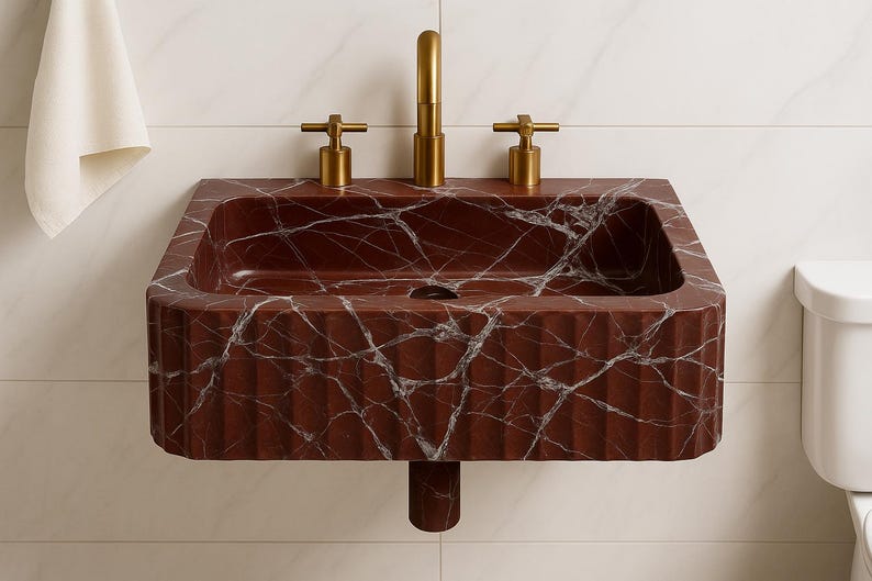 K&ouml;nnte beinhalten: Ein rechteckiges, an der Wand montiertes Waschbecken aus rotem Marmor mit wei&szlig;en Adern. Das Waschbecken hat eine geriffelte Front und goldfarbene Wasserh&auml;hne. Links h&auml;ngt ein wei&szlig;es Handtuch. Breite ca. 60 cm.