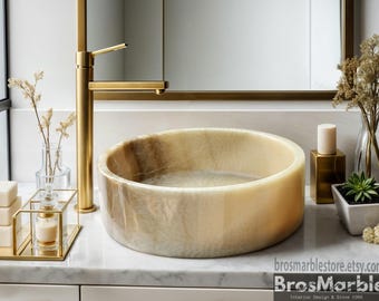 Könnte beinhalten: Ein rundes, cremefarbenes Onyx-Waschbecken mit einem goldfarbenen Wasserhahn. Das Waschbecken steht auf einer weißen Arbeitsplatte mit goldenen Akzenten.
