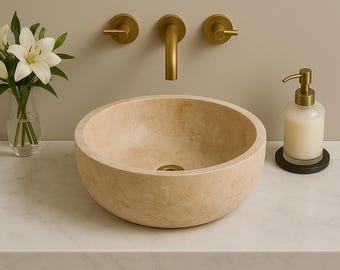 May include: A beige stone vessel sink with a round shape, set on a white countertop. Gold-colored faucet fixtures and a soap dispenser are mounted on the wall. A vase of white flowers is on the left.