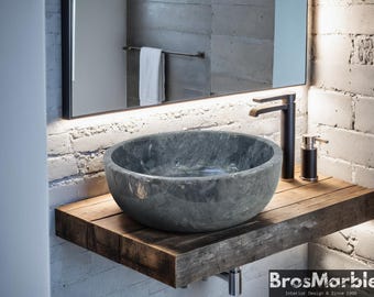 May include: A gray stone vessel sink with a rounded edge sits on a wooden countertop. The sink is in a bathroom with a gray brick wall and a black faucet.