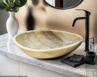 May include: A large, oval-shaped, light brown onyx vessel sink with a white marble countertop. A black faucet and soap dispenser are visible in the background.