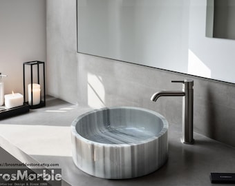 May include: A white marble bathroom sink with a gray veined pattern. The sink is round and sits on a gray countertop. A silver faucet is mounted to the wall above the sink.