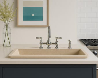 May include: A kitchen sink with a chrome faucet and a beige basin is set into a light-colored countertop. The dark blue cabinets below have gold-colored handles. A framed abstract art piece hangs on the wall, and a white pendant light hangs above.