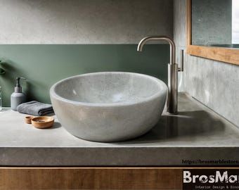 May include: A round, gray stone vessel sink with a brushed nickel faucet. The sink is set on a gray concrete countertop with a wooden cabinet below.