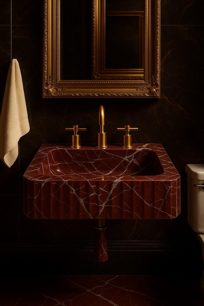 K&ouml;nnte beinhalten: Ein Waschbecken aus rotem Marmor mit goldenen Armaturen. Das Waschbecken hat ein rechteckiges Becken mit geriffelter Front und ist auf einem Sockel montiert. &Uuml;ber dem Waschbecken h&auml;ngt ein goldgerahmter Spiegel.