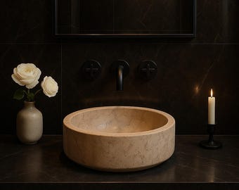 May include: A beige stone vessel sink with a round shape sits on a dark countertop. A black faucet and matching fixtures are mounted on the dark wall. A vase with white roses and a lit candle in a black holder add to the bathroom decor.