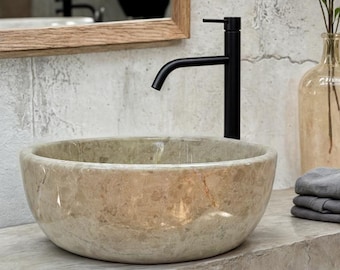 May include: A round, beige marble vessel sink with a black faucet. The sink is on a wooden countertop.