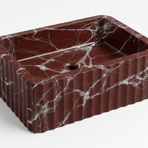 May include: A rectangular bathroom sink made of red marble with white veining. The sink has a fluted exterior design and a drain hole in the center. The overall design is modern and luxurious.