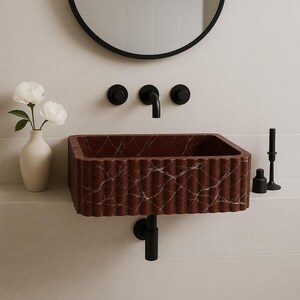 May include: A rectangular, red marble sink with a fluted design, mounted on a white wall. The sink has a black faucet and drainpipe. A black framed mirror hangs above the sink, and a vase with white flowers sits to the left.