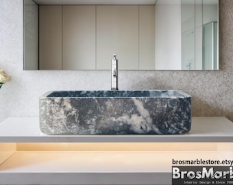 May include: A rectangular, dark gray marble vessel sink with white veining. The sink is mounted on a white countertop with a lit shelf underneath. The sink is in a modern bathroom setting.