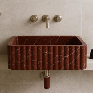K&ouml;nnte beinhalten: Ein rechteckiges Waschbecken aus rotem Marmor mit geriffeltem Design und wei&szlig;en Adern. Das Waschbecken ist an einer Wand mit einem silbernen Wasserhahn und zwei Kn&ouml;pfen befestigt. Eine schwarze Kerze und eine Flasche stehen auf einem Regal.
