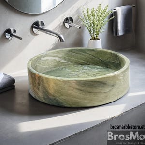May include: A round, green marble sink with a natural, swirling pattern. The sink is set on a gray countertop, with a chrome faucet and a small plant in the background. The sink is a bathroom fixture.