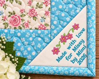 May include: A quilted fabric piece featuring a cream background with pink roses and green leaves, bordered by blue fabric with white floral patterns. A white triangular corner patch displays embroidered pink flowers and the text Made with love for Missy by Mimi 2026.