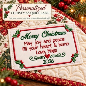 May include: A rectangular fabric quilt label with an off-white background and red border. It features embroidered green holly leaves and red berries. The text reads: Merry Christmas, May joy and peace fill your heart & home, Love, Mags, 2026.