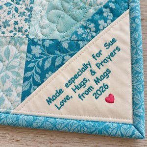 May include: A quilted fabric piece featuring various shades of blue floral and heart patterns. A cream-colored triangular corner patch is embroidered with the text: Made especially for Sue, Love, Hugs, & Prayers from Mags, 2026, and a small pink heart.
